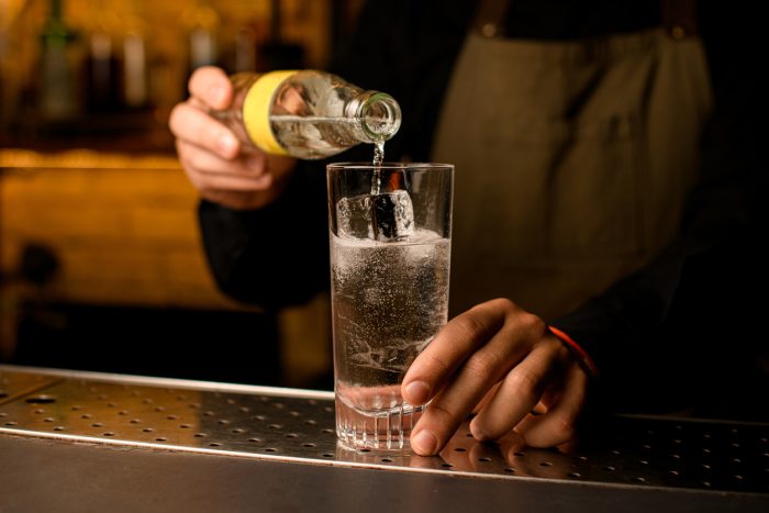 Close-up of glass with cold beverage and ice cubes into which hand of bartender accurate pours fizzy drink from bottle. Cocktail making process.